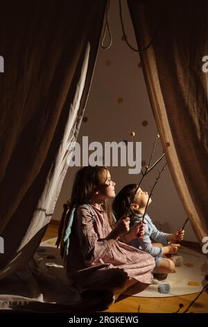 Side view of adorable female kids sitting with folded legs on cotton cloth mat in bright room behind curtain while holding sticks and looking up Stock Photo