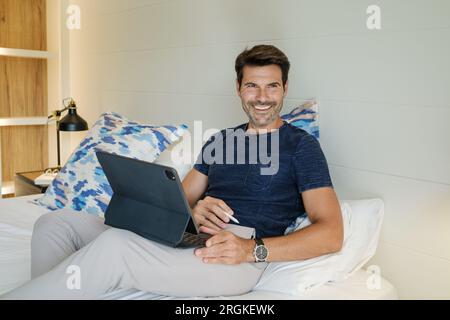 Positive male designer with graphic tablet and stylus sitting on bed and working on creative project at home while looking away Stock Photo
