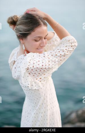 Positive young feminine woman in stylish white dress smiling and looking down while touching head on rocky seashore at sunset Stock Photo