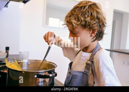 Side view of attentive child in apron with spatula cooking pasta in pot on stove in kitchen Stock Photo