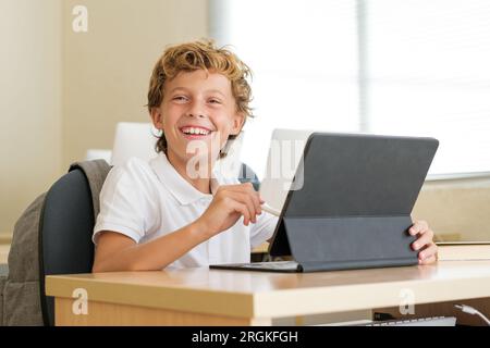 Glad schoolboy using stylus on tablet during lesson while sitting at desk in classroom at school and looking forward Stock Photo