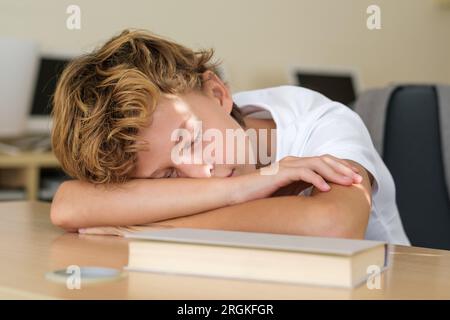Tired schoolboy lying on wooden table with book and sleeping during lesson in classroom at school Stock Photo