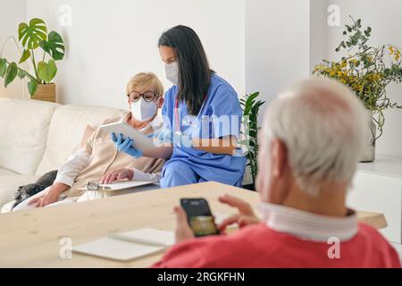 Female physician with tablet talking to senior woman in protective mask against unrecognizable male with smartphone in house room Stock Photo