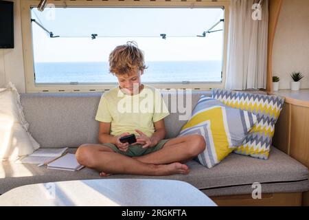 Delighted barefoot boy smiling and browsing social media on cellphone while sitting cross legged on sofa near window in trailer Stock Photo