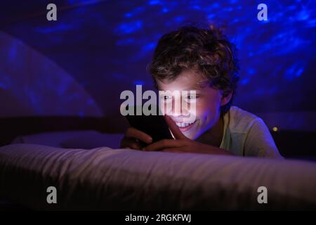 Optimistic boy smiling and texting in social media on mobile phone while lying on bed at night Stock Photo