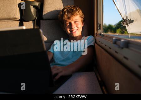 Positive boy looking at camera with smile and using laptop while sitting near camper window on summer weekend day Stock Photo