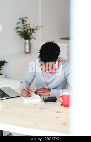 African American man with headset taking notes in copybook at table with calculator and laptop while studying in house Stock Photo