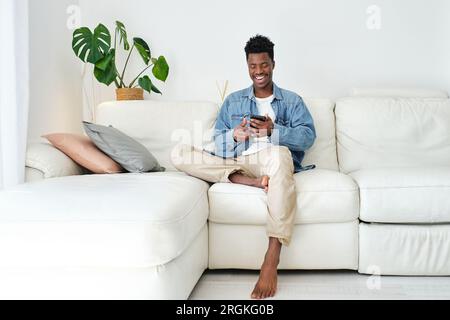 Happy African American male text messaging on cellphone while resting on couch in living room Stock Photo