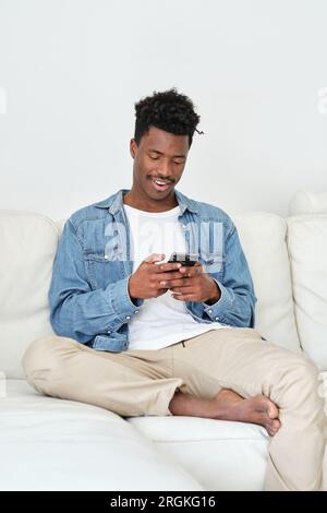 Cheerful young African American male in casual clothes using mobile phone while relaxing on comfy couch in light living room Stock Photo