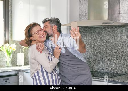 Happy mature man and positive senior woman in aprons hugging and having fun while taking selfie in modern kitchen Stock Photo