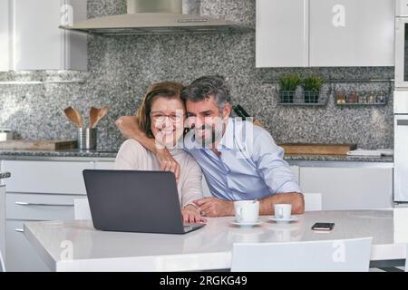 Smiling man hugging elderly woman while looking at laptop screen and laughing in modern marble kitchen at home Stock Photo