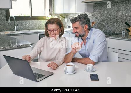 Smiling elderly woman pointing at laptop while sitting with middle aged bearded man at table with cups of drinks in modern marble kitchen at home Stock Photo