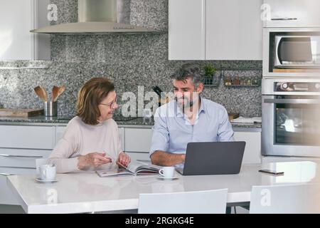 Smiling senior woman reading magazine and looking at mature man typing on laptop while sitting at kitchen table at home Stock Photo