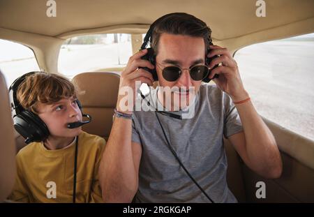 Attentive little boy in casual clothes sitting in four seat aircraft and looking at young male instructor wearing headset before flight Stock Photo