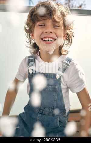 Through glass of positive child with curly hair smiling and looking at camera while doing household routine and washing window on balcony at home Stock Photo