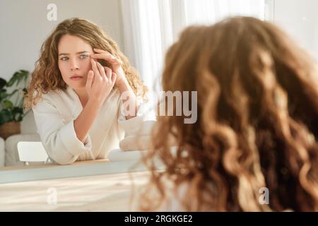 Charming female in bathrobe touching face and looking at wrinkles near eyes in mirror while sitting at table during morning routine at home Stock Photo