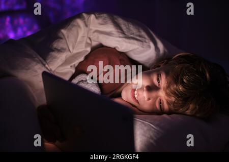 Optimistic child in sleepwear browsing tablet while lying under blanket in bed in dark bedroom at home Stock Photo