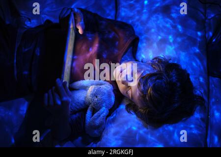 Top view of curly haired kid with toy relaxing on bed and looking at screen while browsing tablet in neon illumination in evening in bedroom Stock Photo