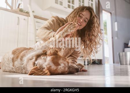 Ground level of smiling young woman with long curly hair giving high five to cute dog while playing together on kitchen floor Stock Photo