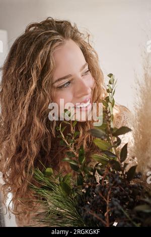 Young woman with long wavy hair looking away happily while standing near green lush foliage in light room Stock Photo