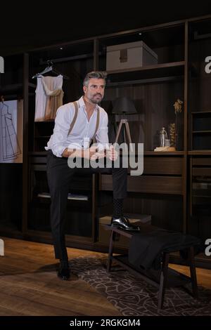 Full body of confident unshaven Hispanic man in elegant clothes looking away and using smartphone while standing against wooden shelves in atelier Stock Photo