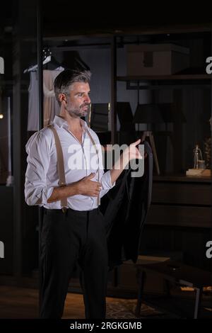 Handsome bearded adult man standing with black jacket in hand in room with black walls Stock Photo