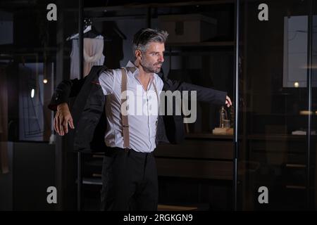 Senior self confident man in suit standing near glass wall and putting on black jacket with hands to sides Stock Photo