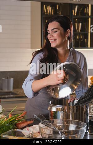 Smiling young woman in apron having fun while cooking healthy dinner in cozy kitchen at home Stock Photo