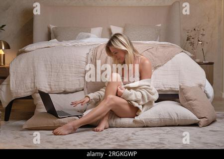 Full body of barefoot woman in nightwear sitting on pillows on floor and working on netbook in cozy bedroom at home Stock Photo