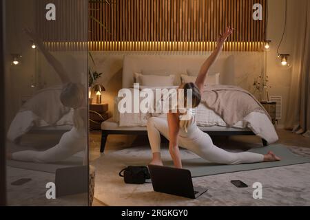 Side view of young slim woman in activewear practicing yoga in Anjaneyasana and stretching arm while watching online tutorial on laptop in bedroom Stock Photo