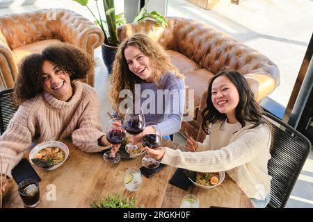 From above of positive young multiracial girlfriends sitting at wooden table with glasses of red wine and bowls with ramen Stock Photo