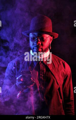 Bearded black man in hat standing in studio with smoke and using smartphone on black background with red and blue neon illumination Stock Photo