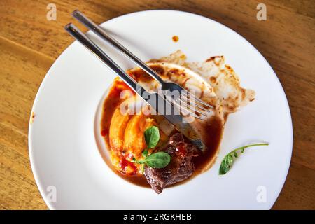 Forks on a plate with leftover food from a dish of veal cheeks with coconut mojo and tuna puree with orange sauce served in a white ceramic plate in a Stock Photo