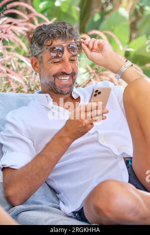 Positive bearded man in casual clothes and eyeglasses sitting on bench while browsing smartphone and smiling happily Stock Photo