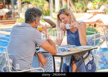 Cheerful woman having cup of coffee while spending time with man drinking juice at table in resort near pool on sunny day Stock Photo