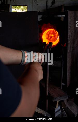 Crop anonymous male glass blower putting piece of glass in melting oven while standing on distance in workshop Stock Photo