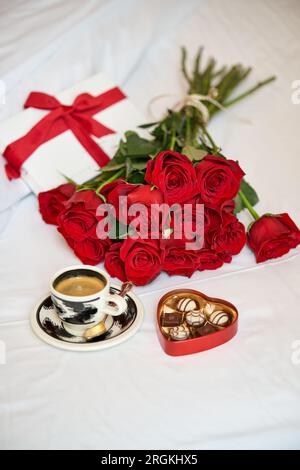 From above of composition of romantic gift consisting of bouquet of red roses cup of fresh coffee and heart shaped box of chocolates placed of bed in Stock Photo