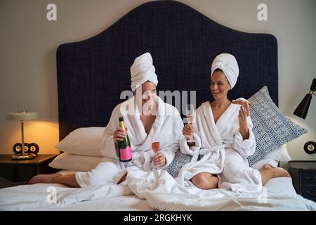 Cheerful female friends resting on bed in terry bathrobes drinking champagne and eating delicious croissants Stock Photo