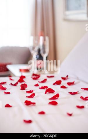 Closeup of comfortable bed with scattered red roses petals and bottle of champagne with glasses placed on bedside table in hotel room prepared for hon Stock Photo