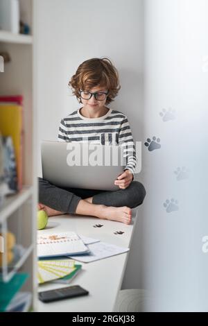 Focused preteen smart boy in glasses browsing social media on netbook and looking at screen while spending time at home Stock Photo