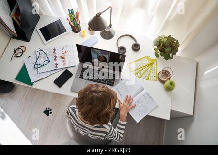 From above of schoolboy with exercise book doing homework during video chat with classmate on laptop at table with notebooks Stock Photo