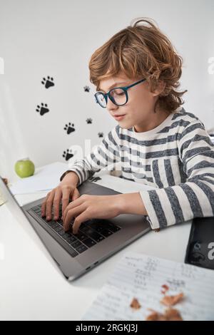 Focused clever curly haired boy surfing internet via netbook and doing assignment while studying remotely at home Stock Photo