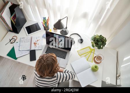 From above of anonymous kid having video call on laptop with classmate while doing schoolwork at table with exercise book Stock Photo