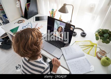 From above of diligent kid making video chat with classmate on laptop at table with exercise book while doing homework together Stock Photo