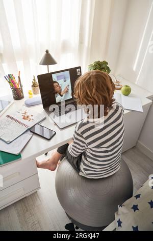 From above of unrecognizable boy video chatting with classmate via laptop while doing homework together at table in light room Stock Photo