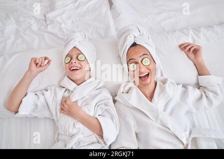 Top view of laughing woman and boy with cucumber slices on eyes wearing white bathrobes and towel turbans resting on soft bed in bedroom at home Stock Photo
