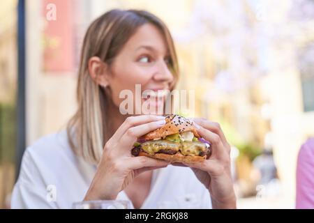 Positive young woman with loose hair in white blouse enjoying appetizing succulent burger while having lunch at outdoor cafe Stock Photo