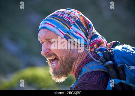 Portrait of bearded middle aged male hiker with wrinkles wearing multicolored headscarf and backpack standing near blurred picturesque landscape and y Stock Photo