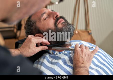 Anonymous barber cutting hair of bearded man with scissors and comb while working in hairdressing salon Stock Photo