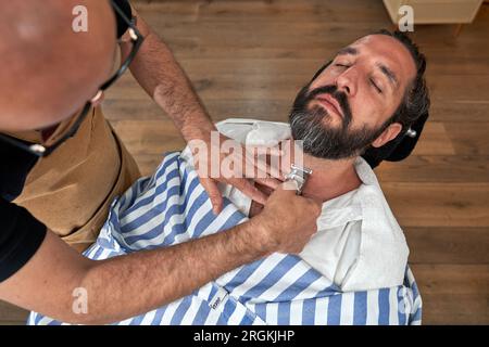 From above of crop anonymous male barber using trimmer while working with bearded client in hairdressing salon Stock Photo